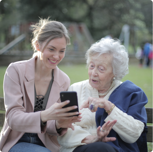 An elderly woman sitting with her granddaughter on a bench and pointing at the phone screen